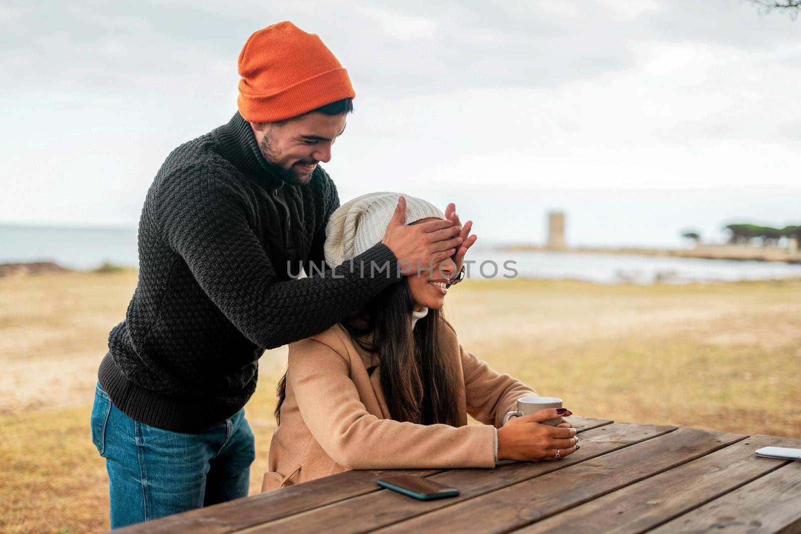 Young heterosexual couple having fun outdoor in sea resort wearing fall-winter clothes and wool hats - Handsome smiling guy blindfolds from behind with hands his girlfriend sitting with a tea cup by robbyfontanesi
