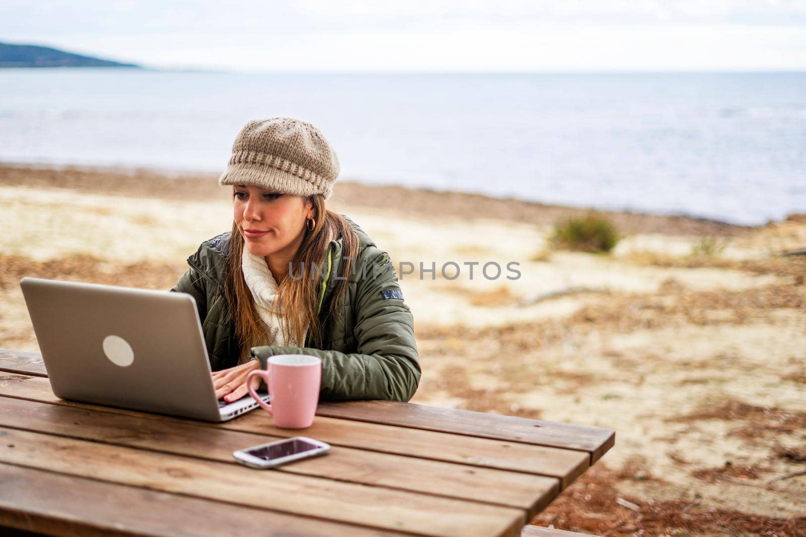 Internet female freelance working outdoor in sea resort sitting at wooden table wearing fall-winter clothes and wool hat with tea cup close to her aluminum laptop - Change your life making online job by robbyfontanesi