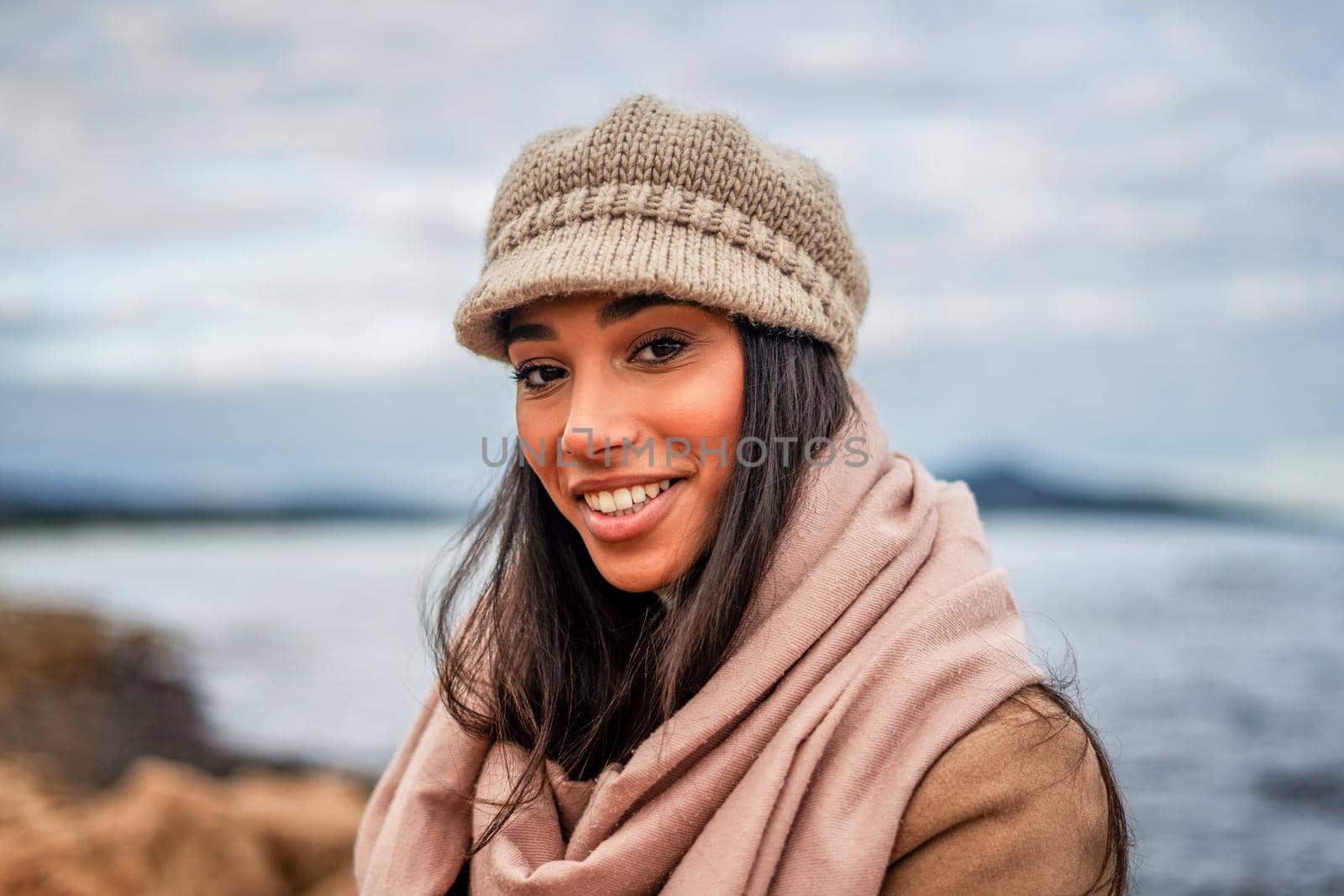 Cute Hispanic young woman with long black hair looking at camera posing outdoor in sea resort with big pink scarf, beige wool hat, and toothy smile - Beautiful female model head and shoulders portrait