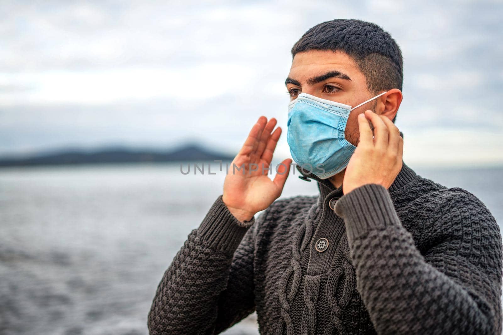 Handsome guy in dark wool sweater outdoor in sea resort has just worn face medical protection mask against Coronavirus pandemic virus - Young man living winter vacation with Covid-19 social disease