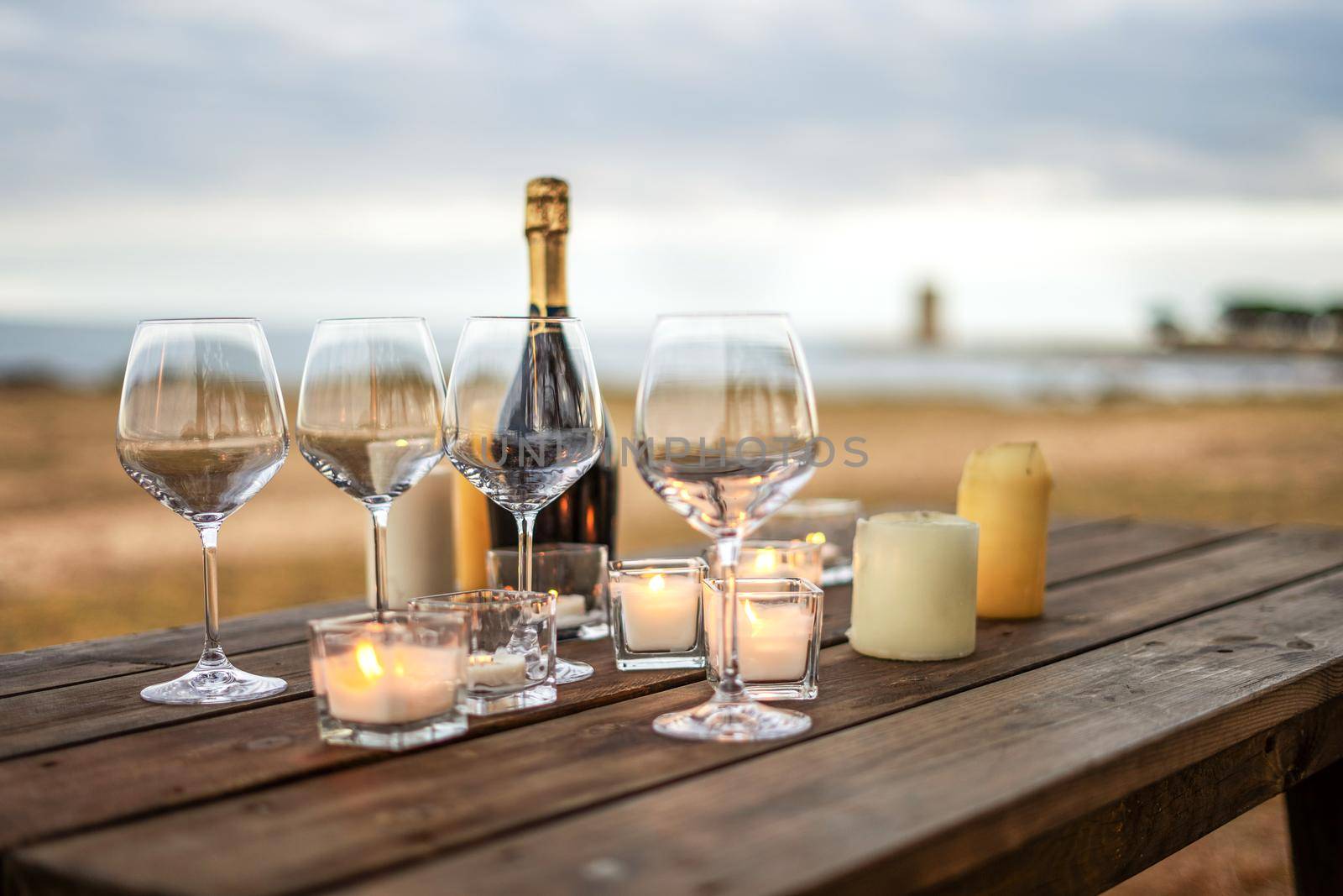 All is ready for the end of year toast in winter holidays by the sea - Celebration scene with selective focus on empty glasses, closed champagne bottle, and candles on a wooden table of outdoor bar
