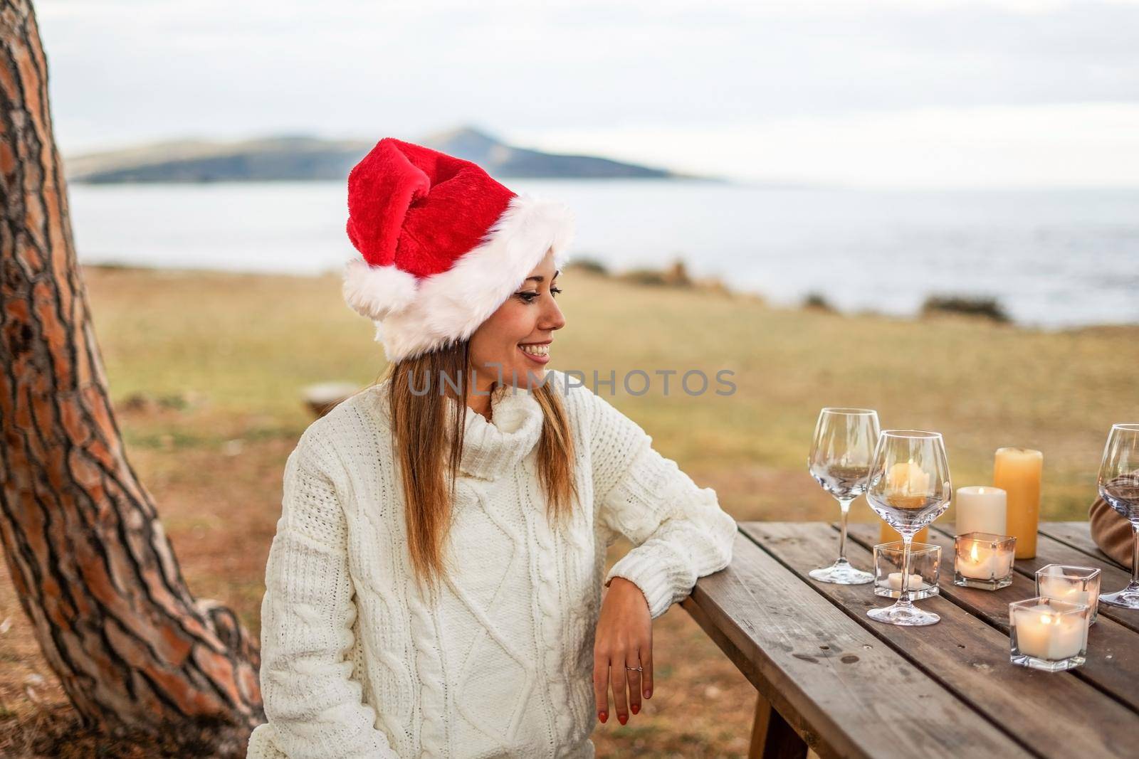 Beautiful young woman smiling sitting outdoor at a wooden table posing with Santa Klaus hat and white wool sweater watching empty glasses before toast for celebrating holidays in sea winter vacation by robbyfontanesi