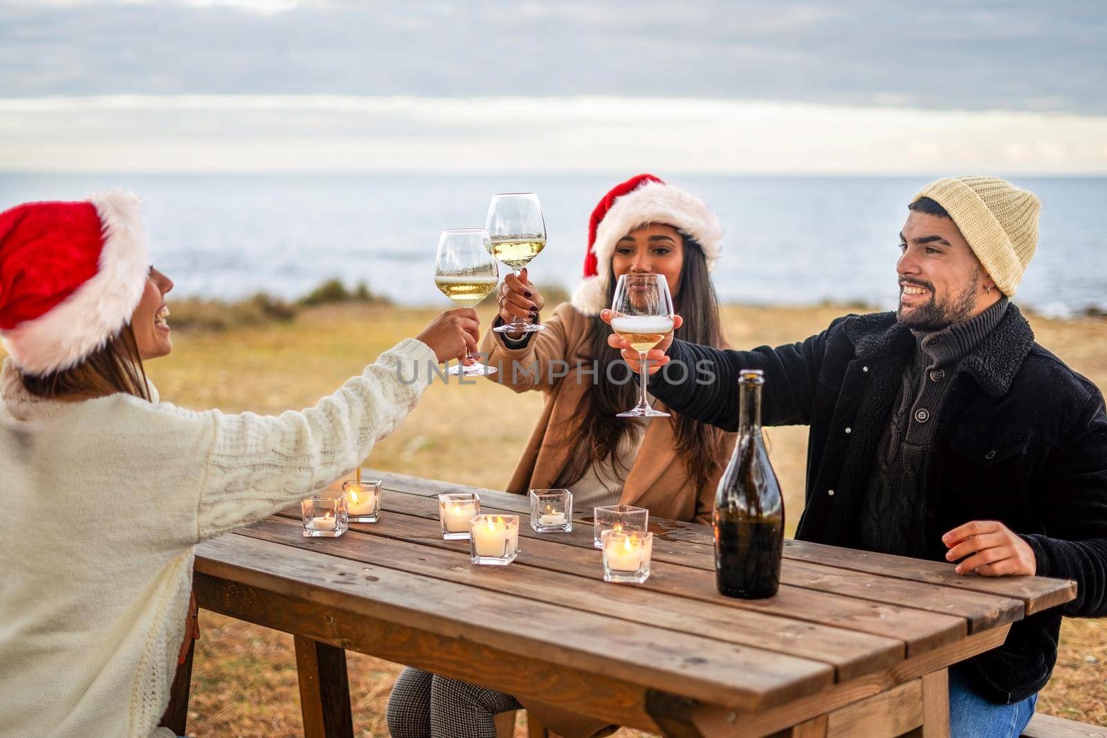 Group of young mixed race friends toasting outdoor sitting at a wooden table near the water of the ocean in sea winter holidays - Multiracial people celebrating in the nature Christmas and New Year by robbyfontanesi