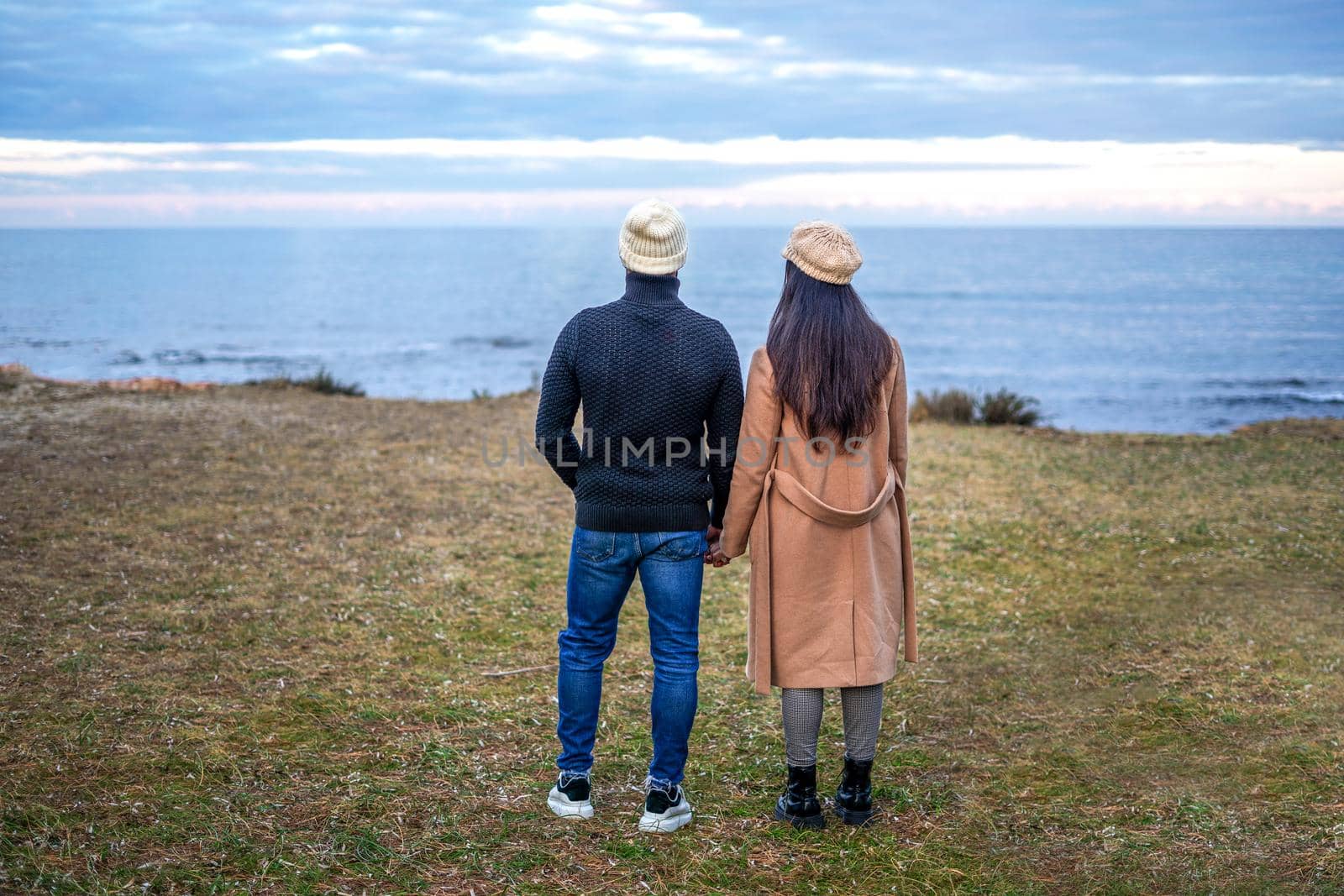 Romance scene of young heterosexual couple view from back holding hands watching the horizon on the sea - Two unrecognizable people wearing fall-winter clothes looking the ocean sunset with cloudy sky