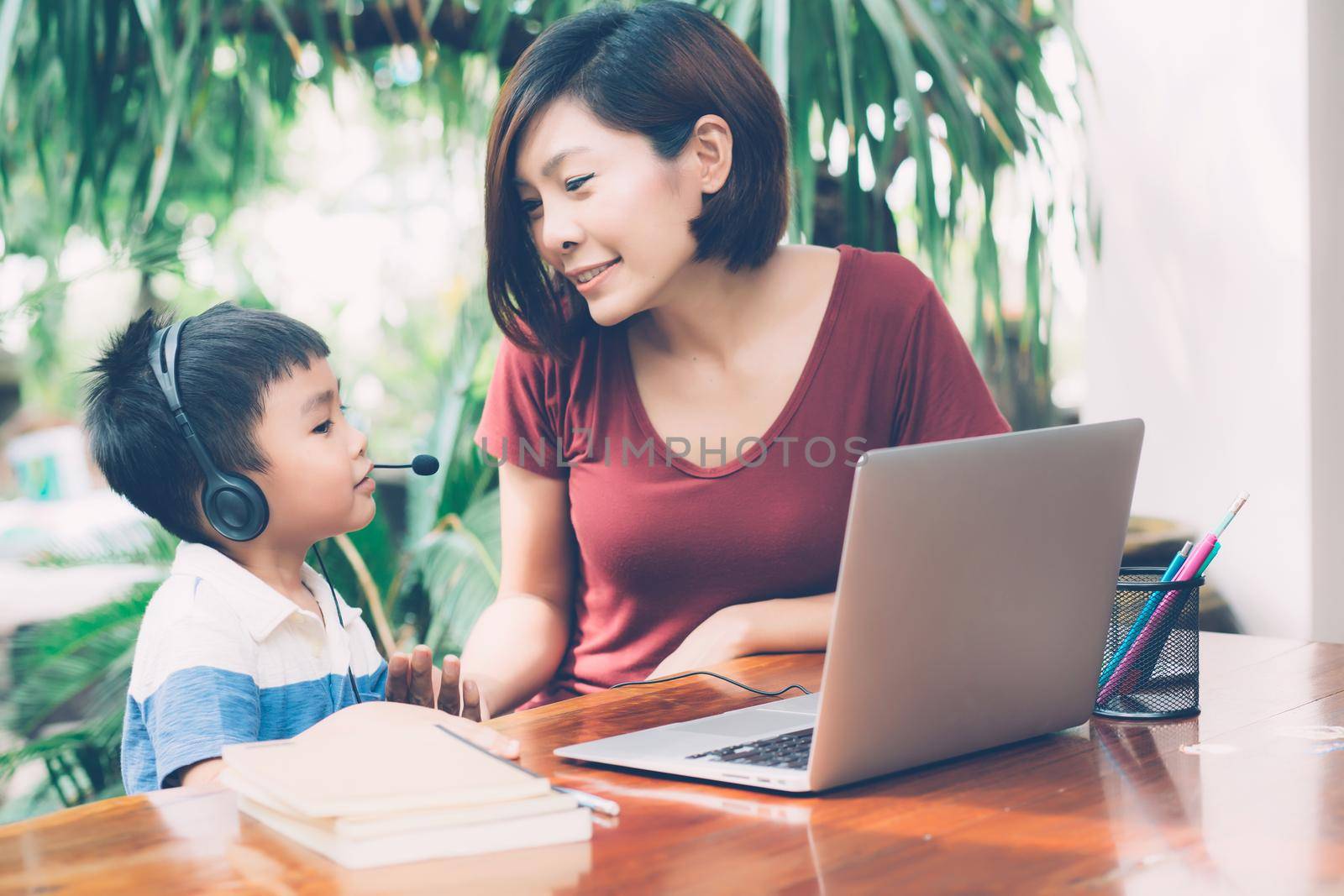 Young asian mother and son using laptop computer for study and learning together at home, boy wearing headphone for e-learning with distancing, teacher or mom support child, education concept.