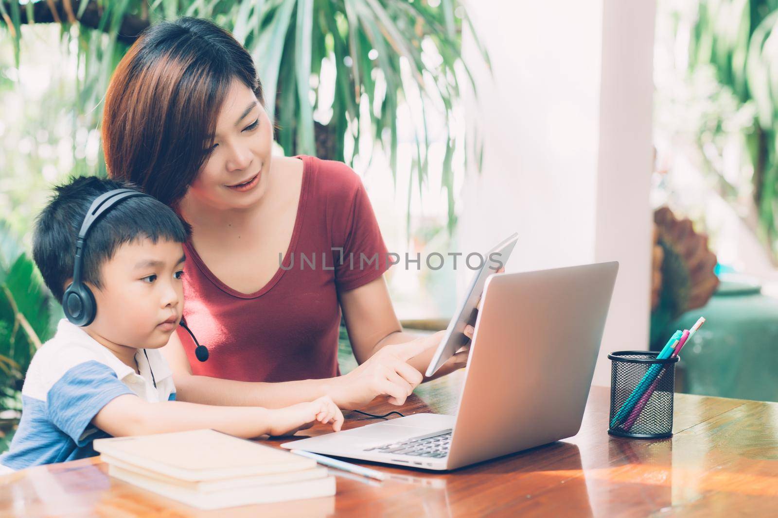 Young asian mother and son using laptop computer and tablet for study and learn together at home, boy wearing headphone for e-learning with distancing, teacher or mom support child, education concept. by nnudoo
