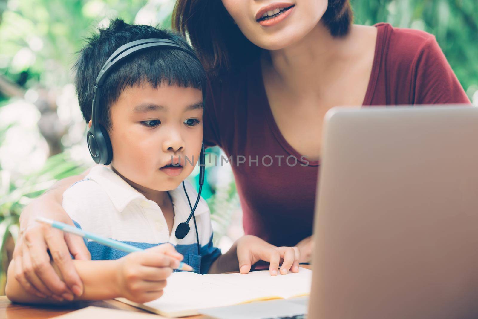 Young asian mother and son using laptop computer for study and learning together at home, boy writing on notebook for homework and wearing headphone, teacher or mom support child, education concept. by nnudoo