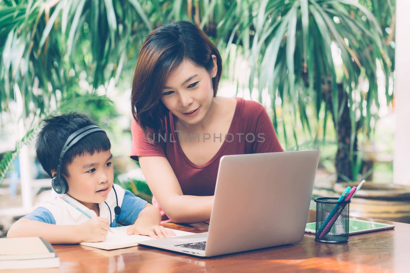 Young asian mother and son using laptop computer for study and learning together at home, boy writing on notebook for homework and wearing headphone, teacher or mom support child, education concept.