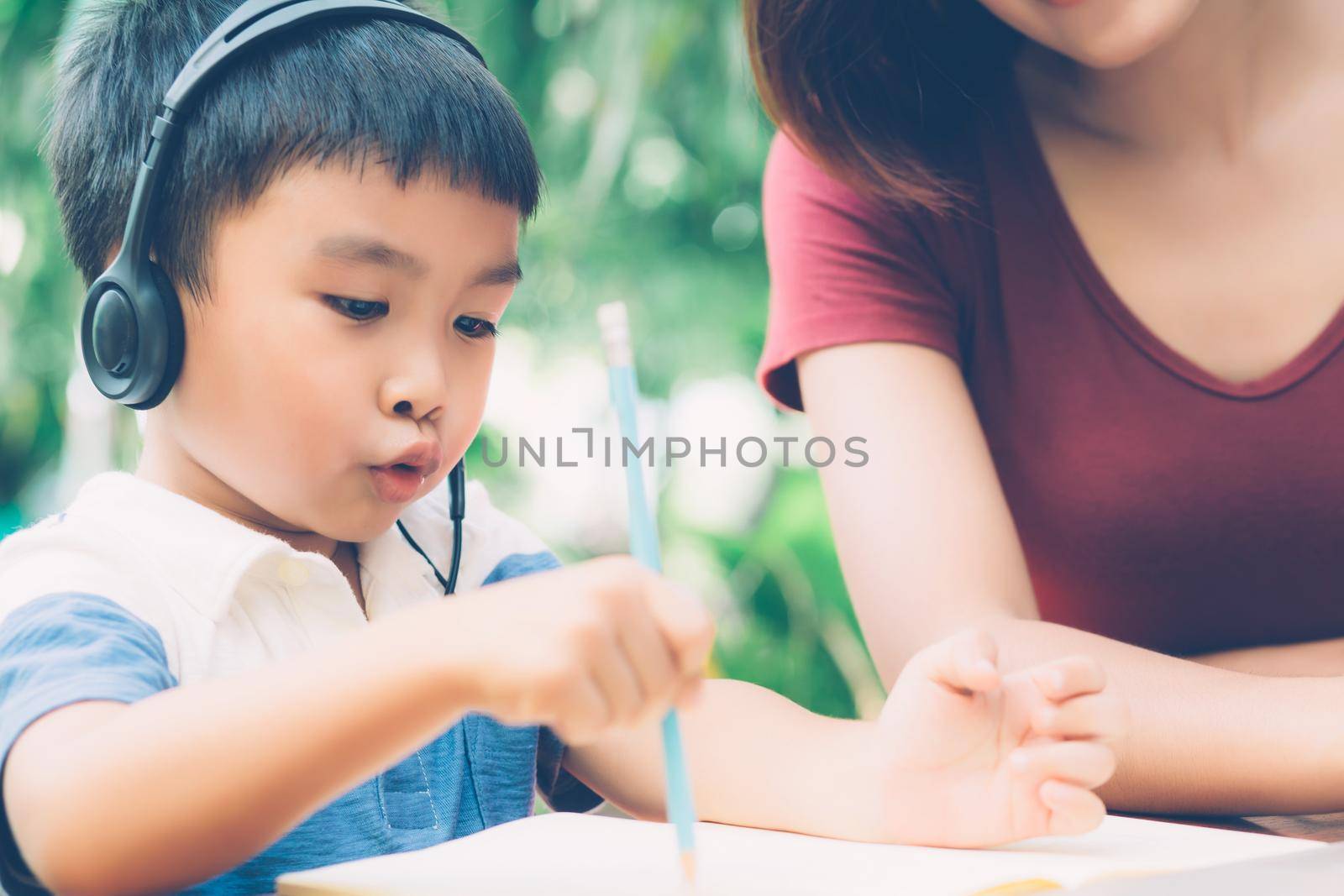 Young asian mother and son using laptop computer for study and learning together at home, boy writing on notebook for homework and wearing headphone, teacher or mom support child, education concept.