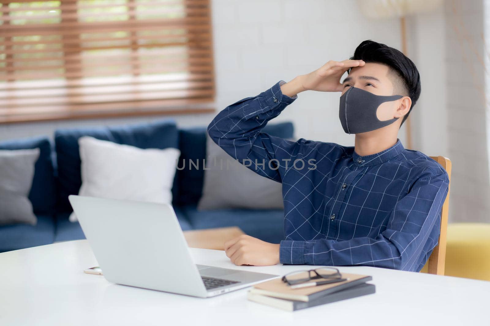 Young business man in face mask working from home with laptop computer and bored on desk for protection for outbreak of covid-19, male quarantine stay home using notebook with unhappy, new normal.