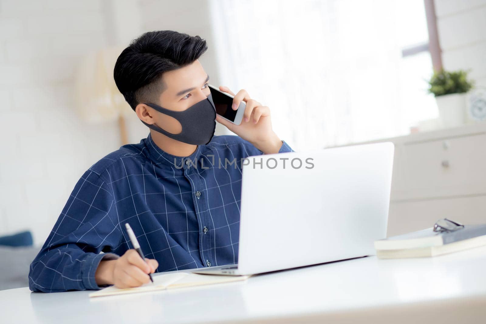 Young asian business man in face mask talking smartphone and work on laptop computer for protect covid-19, businessman quarantine work from home and writing notebook, social distancing and stay home.
