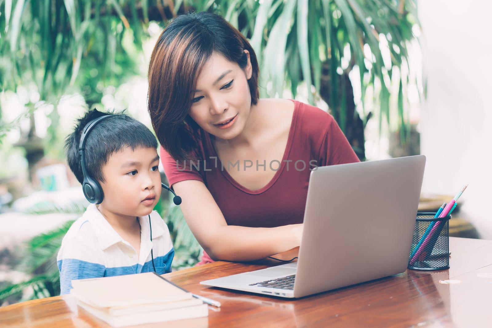 Young asian mother and son using laptop computer for study and learning together at home, boy wearing headphone for e-learning with distancing, teacher or mom support child, education concept.