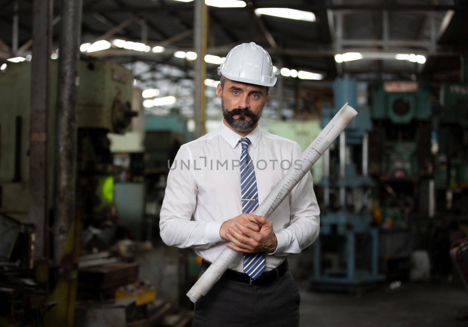 The bearded hipster director with engineer hat standing against machine in production in factory