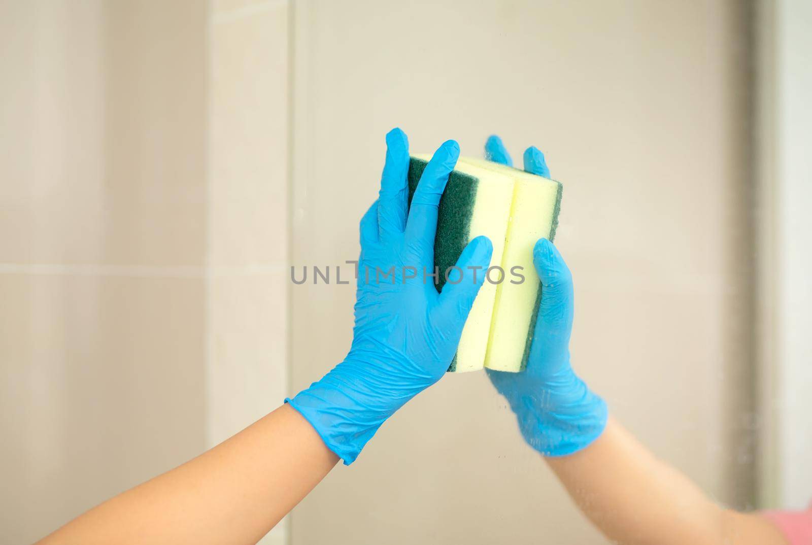 Close-up women hand in a blue rubber glove and cleaning spong in the picture, removes and washes bathroom sink and mirror.