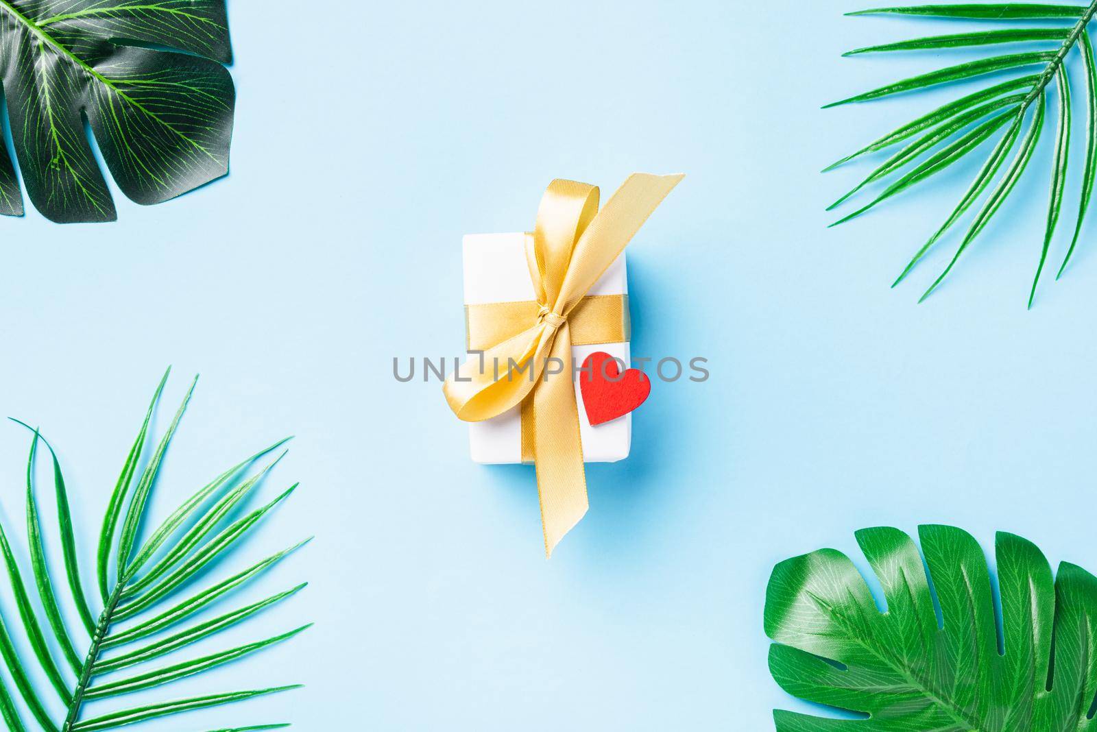 Valentines' day concept. White gift box postcard with a gold bow ribbon and Tropical jungle Monstera green leaves and red heart isolated on blue background with copy space, Symbol of love. Top View
