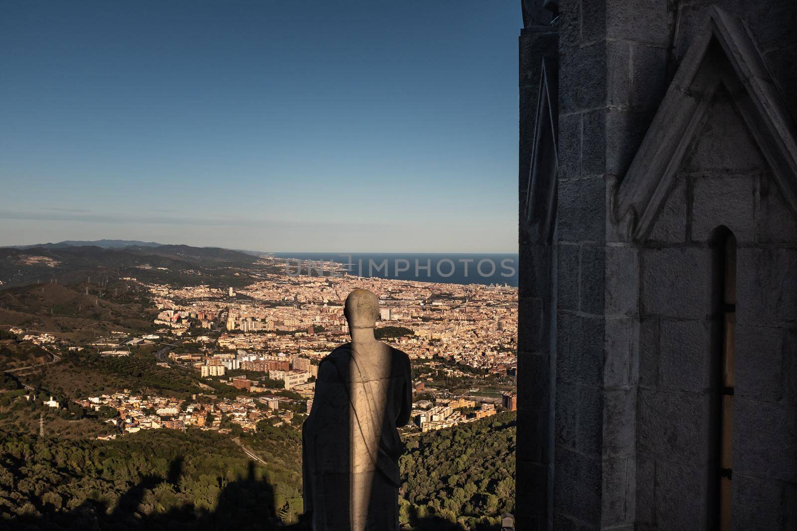 View over northern Barcelona (Spain) from the Temple del Sagrad Cor (Catalan for Temple Sacred Heart of Jesus). A sculpture of an apostle partially illuminated by the sun. by Riccarduska