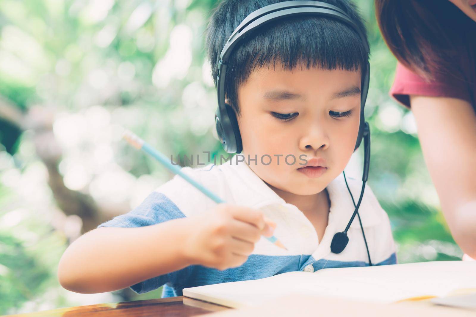 Young asian mother and son using laptop computer for study and learning together at home, boy writing on notebook for homework and wearing headphone, teacher or mom support child, education concept.