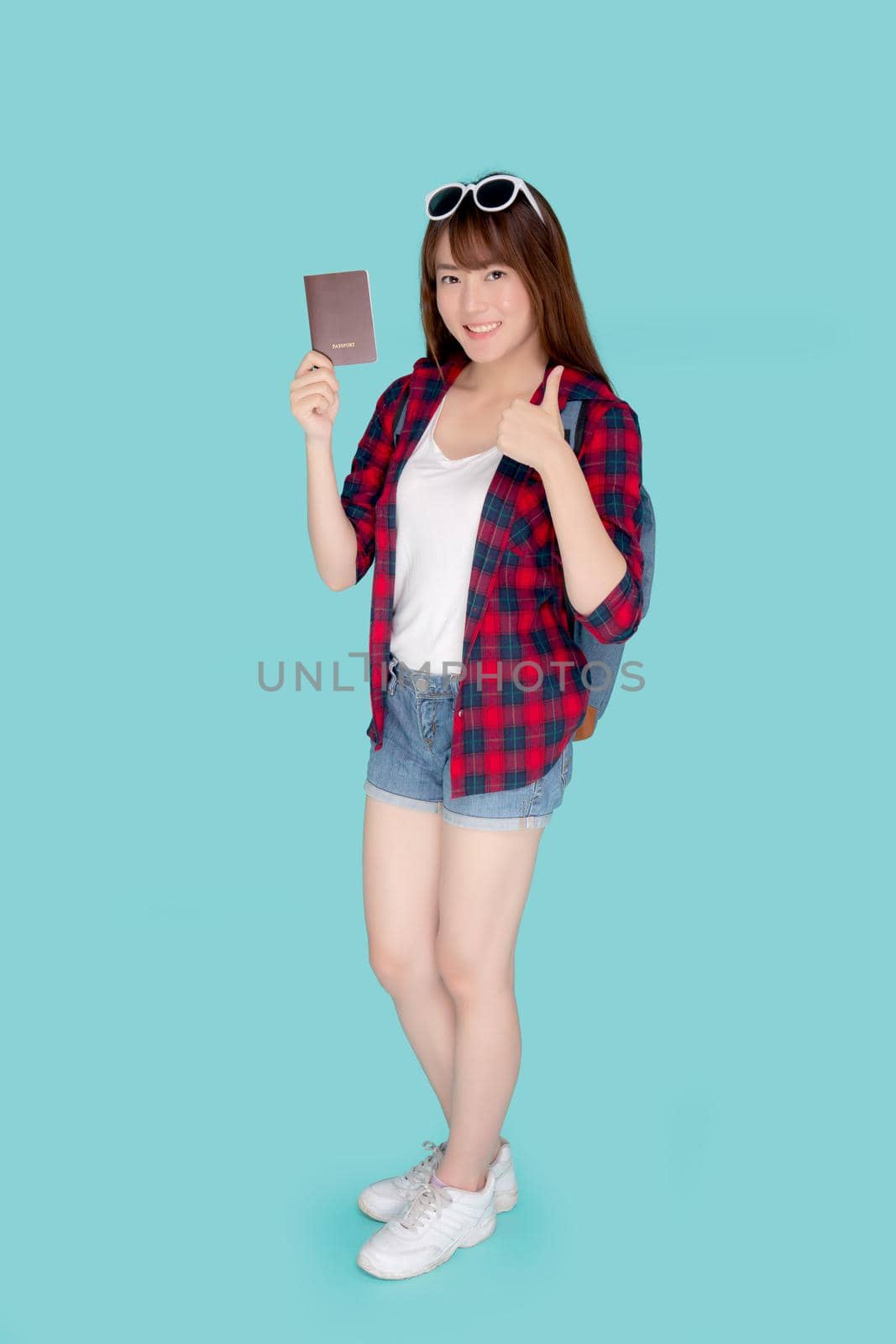 Beautiful portrait young asian woman wear sunglasses on head and backpack smiling gesture thumbs up enjoy summer travel holiday isolated blue background, asia girl fashion trip and holding passport.