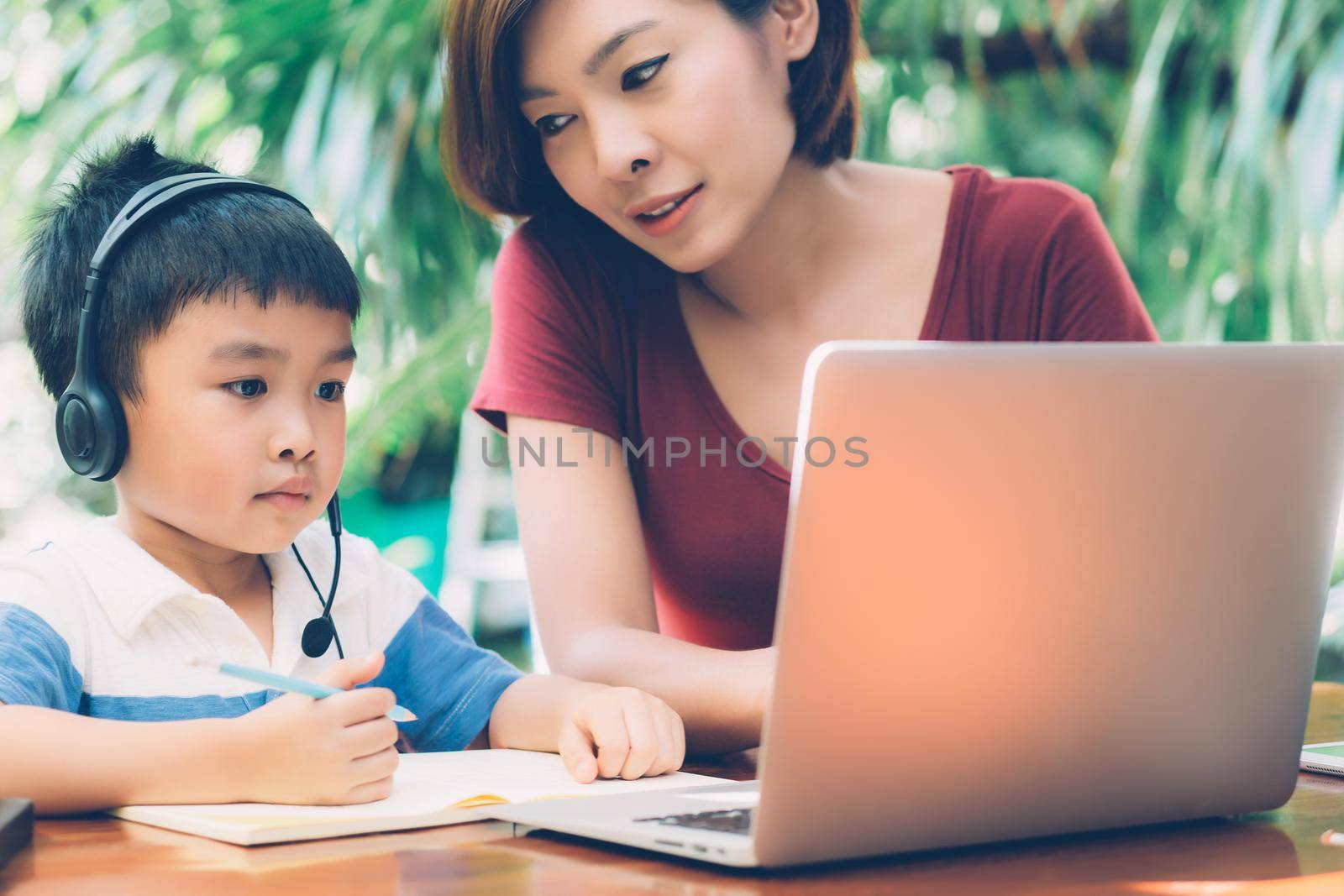 Young asian mother and son using laptop computer for study and learning together at home, boy writing on notebook for homework and wearing headphone, teacher or mom support child, education concept. by nnudoo