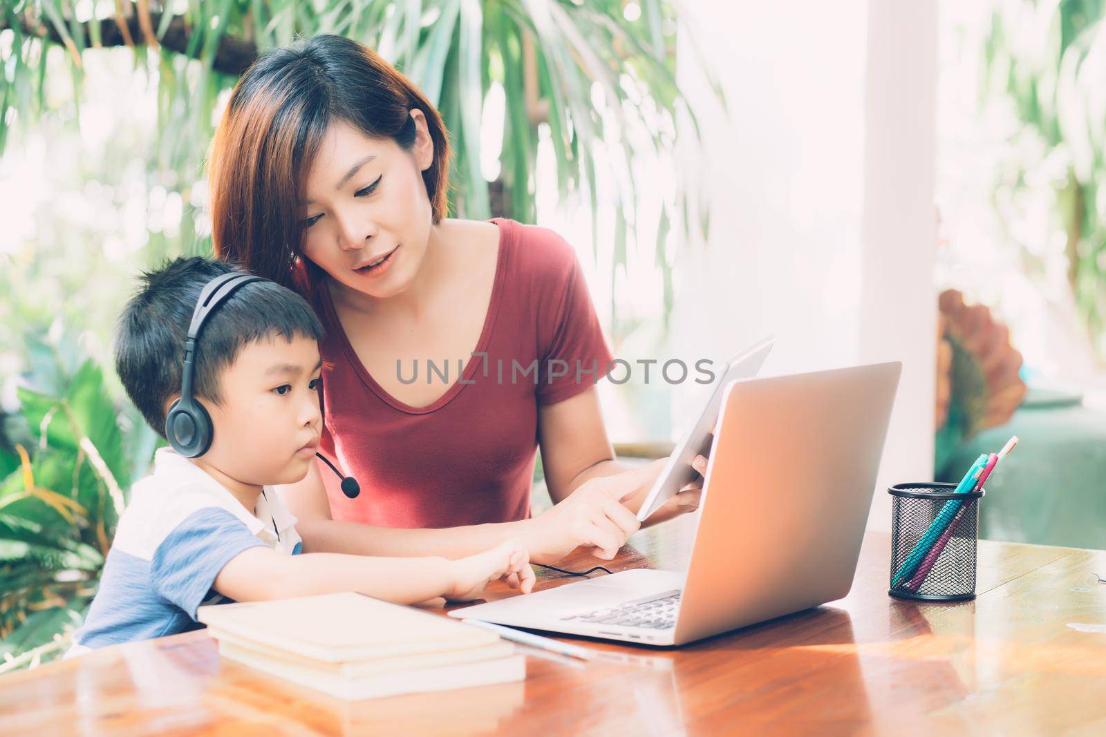 Young asian mother and son using laptop computer and tablet for study and learn together at home, boy wearing headphone for e-learning with distancing, teacher or mom support child, education concept. by nnudoo