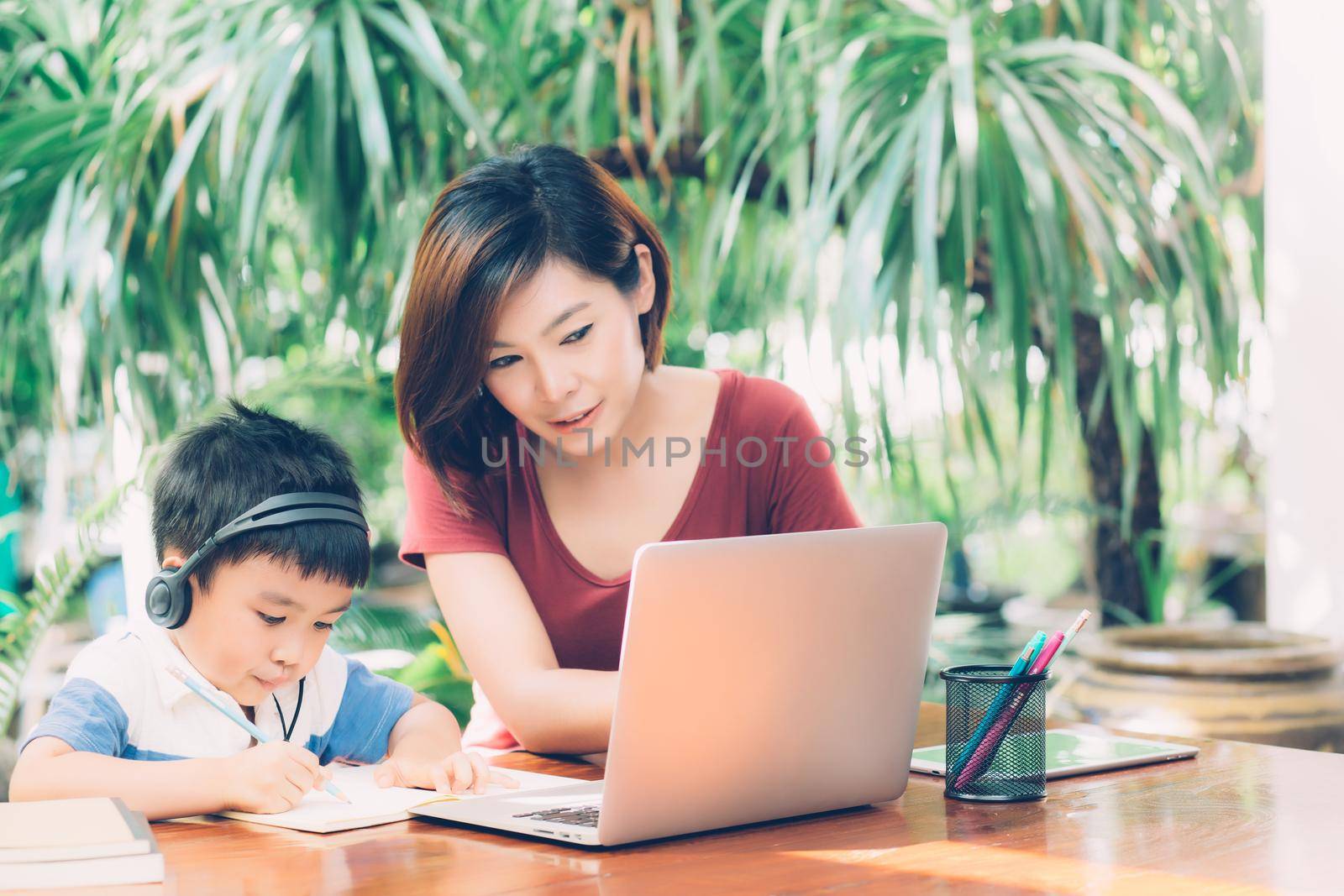 Young asian mother and son using laptop computer for study and learning together at home, boy writing on notebook for homework and wearing headphone, teacher or mom support child, education concept. by nnudoo