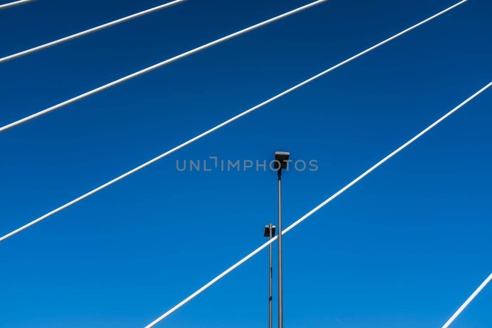 Pescara, Italy: white steel cables of a suspension bridge intersect with street lights. Abstract composition against a bright blue sky.