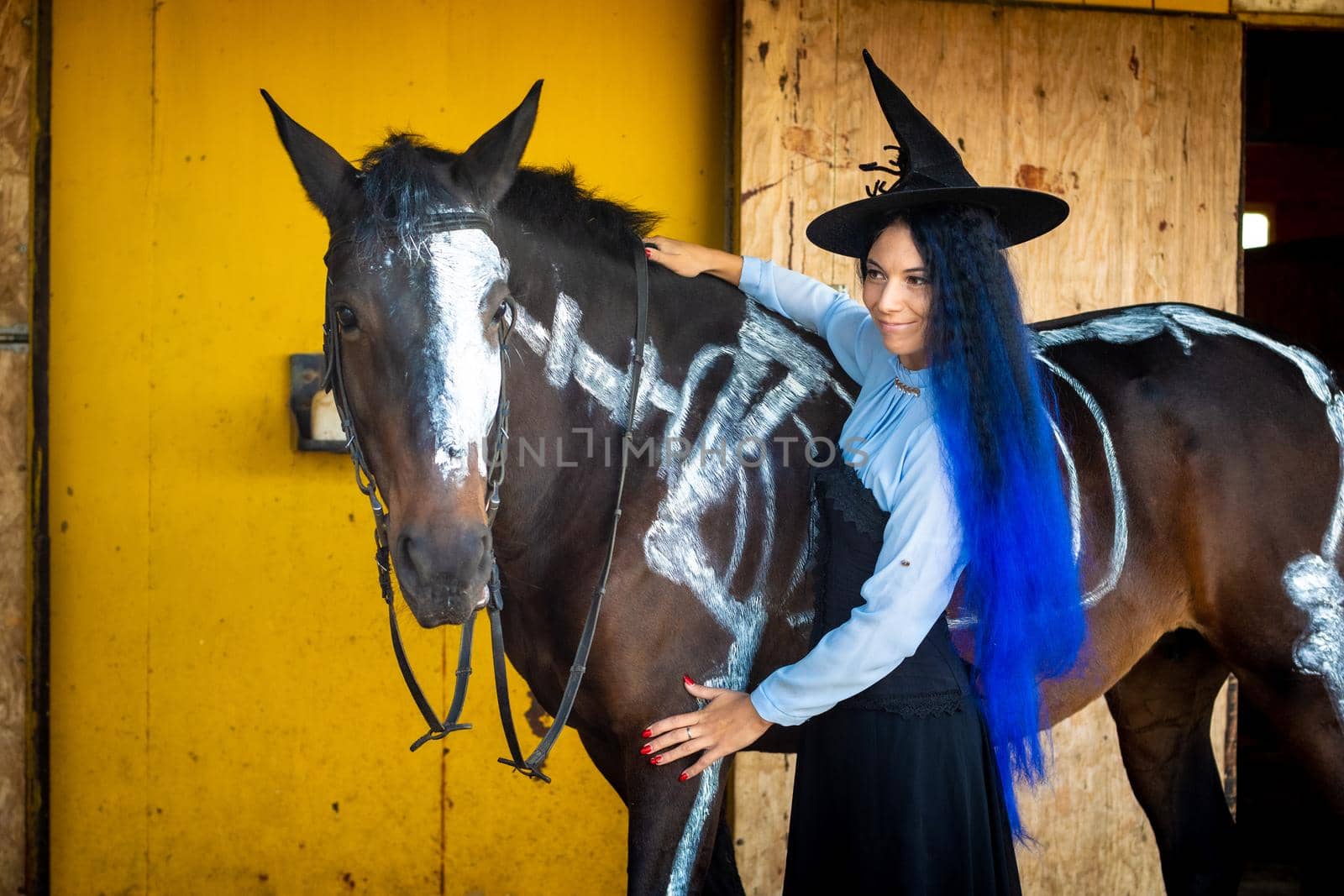A girl dressed as a witch stands by a horse on which a skeleton is painted in white paint