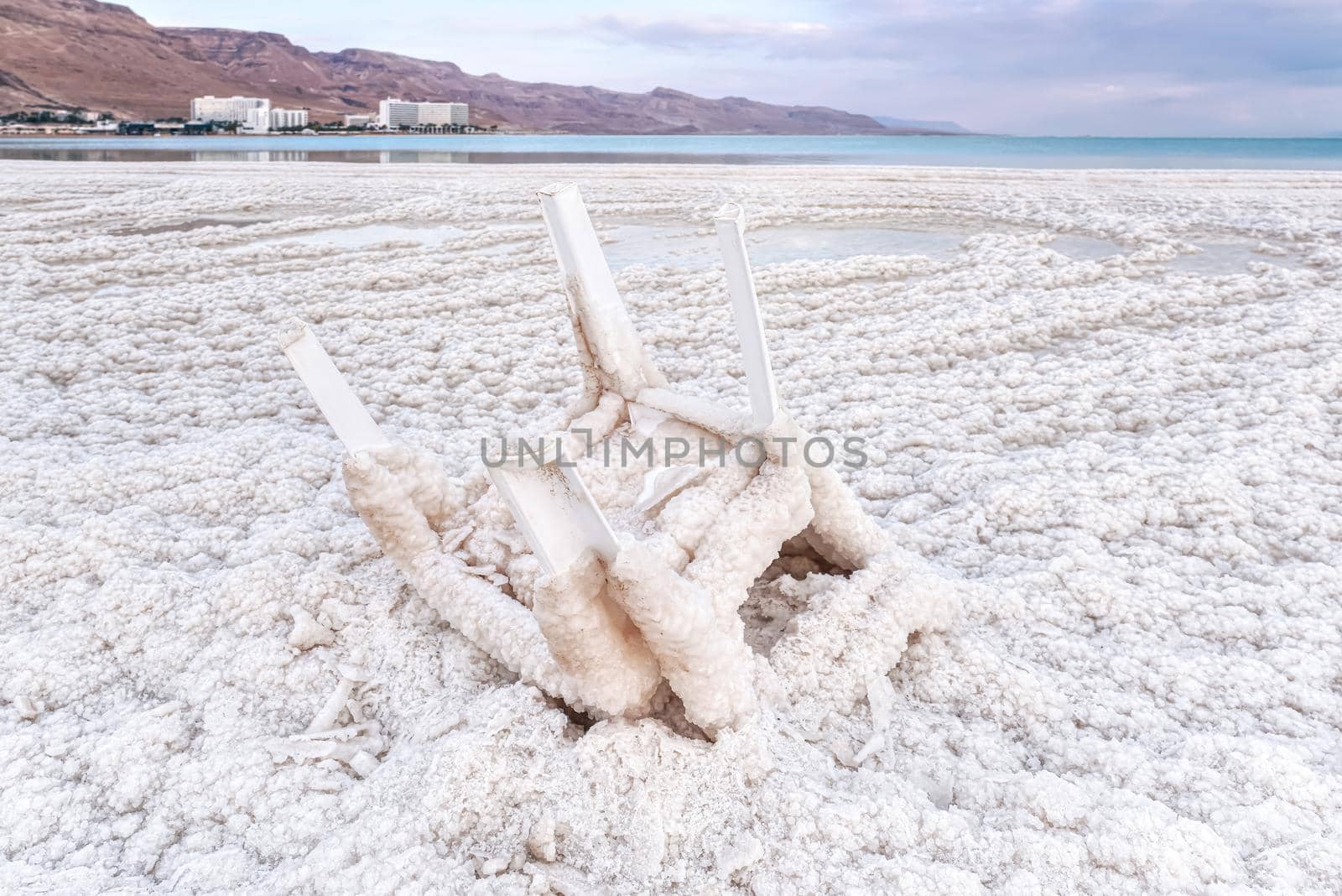 Small plastic chair completely covered with crystalline salt on shore of dead sea, closeup detail, blurred hotel resorts in distance - Ein Bokek, Israel.