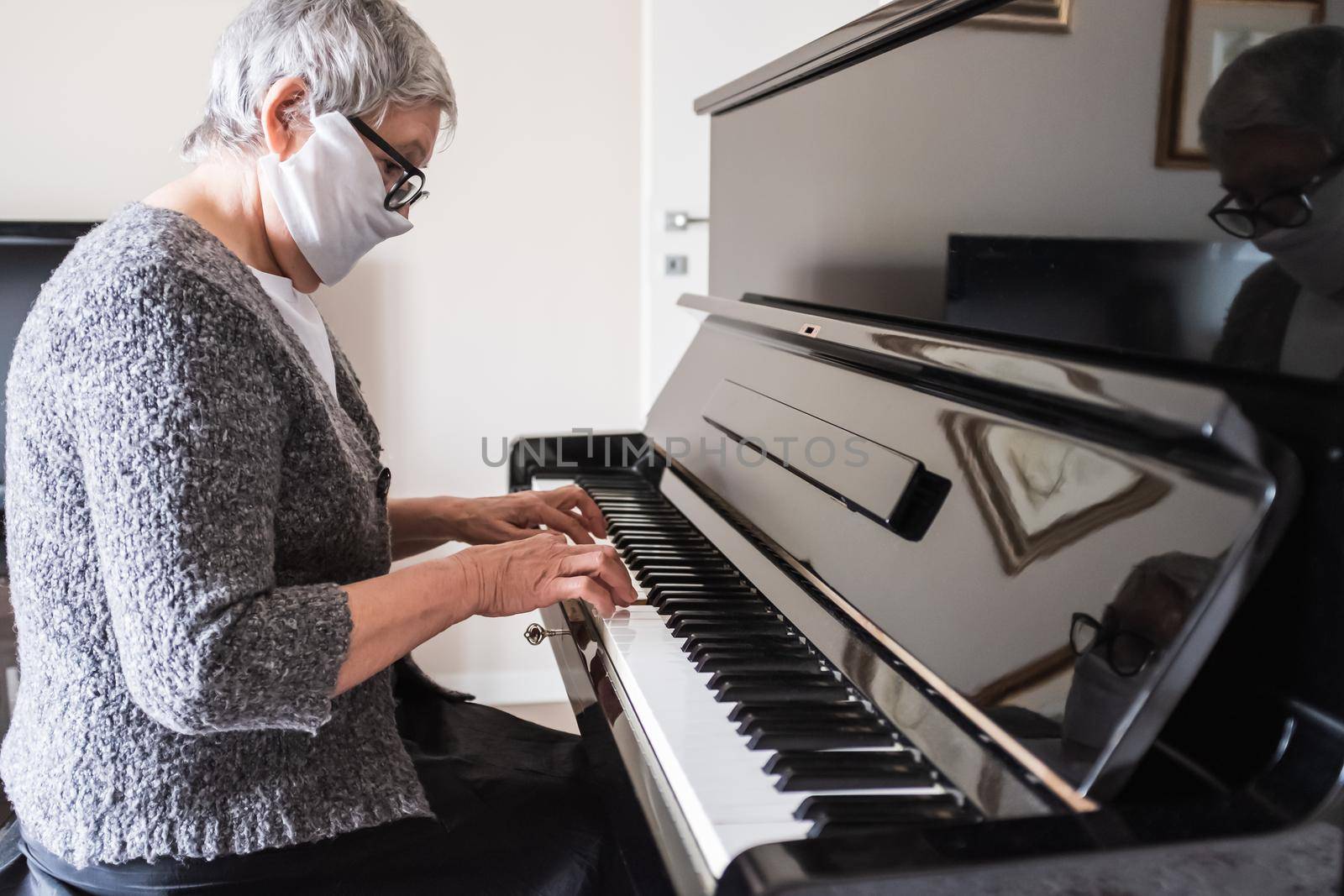 Side view portrait of a senior Italian woman with glasses and face mask playing the piano at home. Prevention for COVID-19 coronavirus pandemic and indoor activity during isolation. Concept of resilience.
