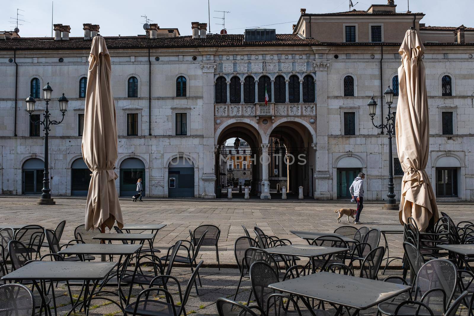 Deserted tables and chairs outside a restaurant. Only two persons walking across the main town square in Brescia during the coronavirus emergency lockdown.