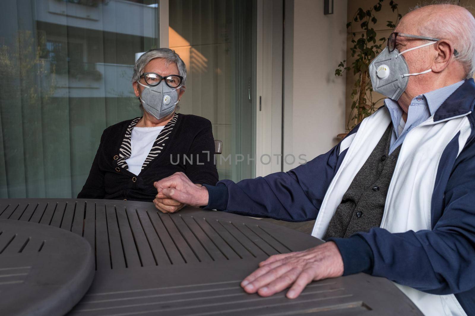 An elderly couple wearing a protective mask and holding hands. They are sitting at terrace of their house in Brescia, Lombardy, Italy. by Riccarduska