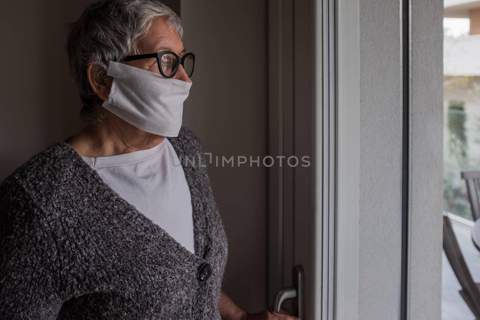 Side view of an old lady standing next to the window of her house. Contagion control measures urge the elderly population to stay at home so as to avoid contact with other people.