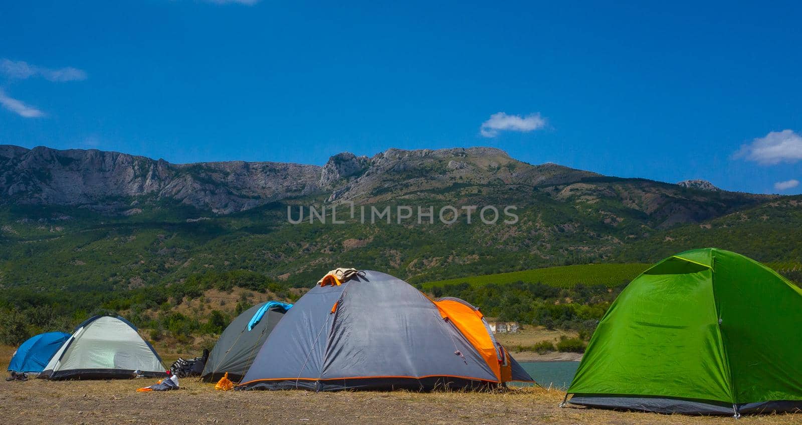 Multicolored tents on the shore of a mountain lake against the backdrop of inaccessible rocks surrounded by a green forest.