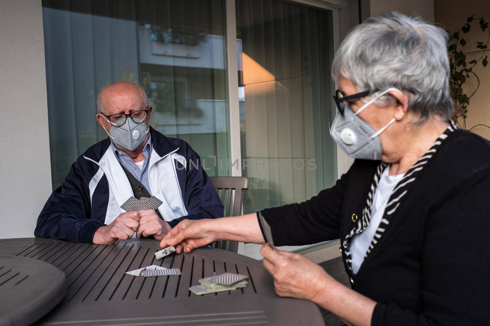 Portrait of seniors couple wearing a protective mask playing card game while sitting at outdoor table of their house in Brescia, Lombardy, Italy. by Riccarduska