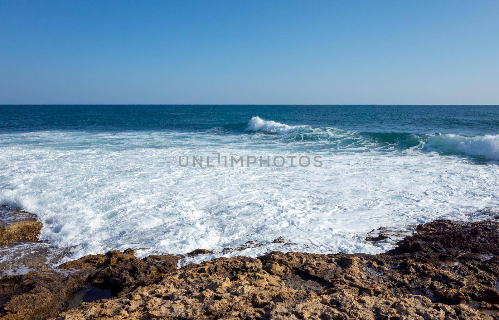 Waves break on the rocky shore, forming mountains of white foam on a clear Sunny day.
