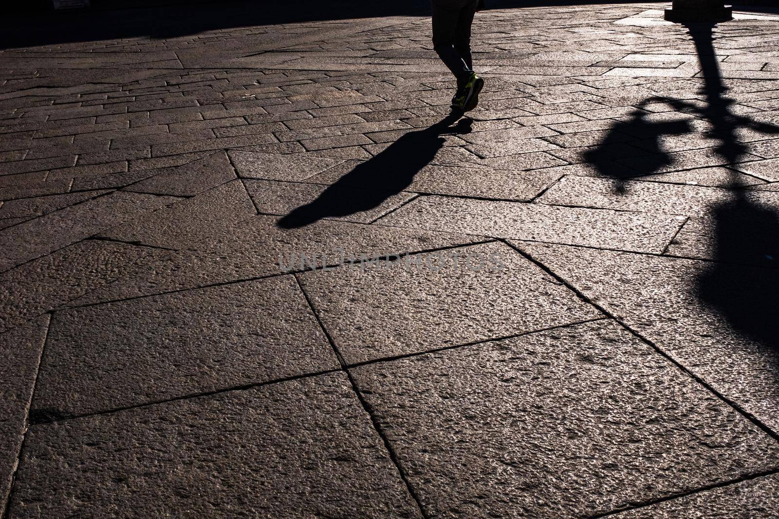 Silhouette of a man and a shadow of a street light on a paving stone in the old town square in Brescia (Italy). by Riccarduska