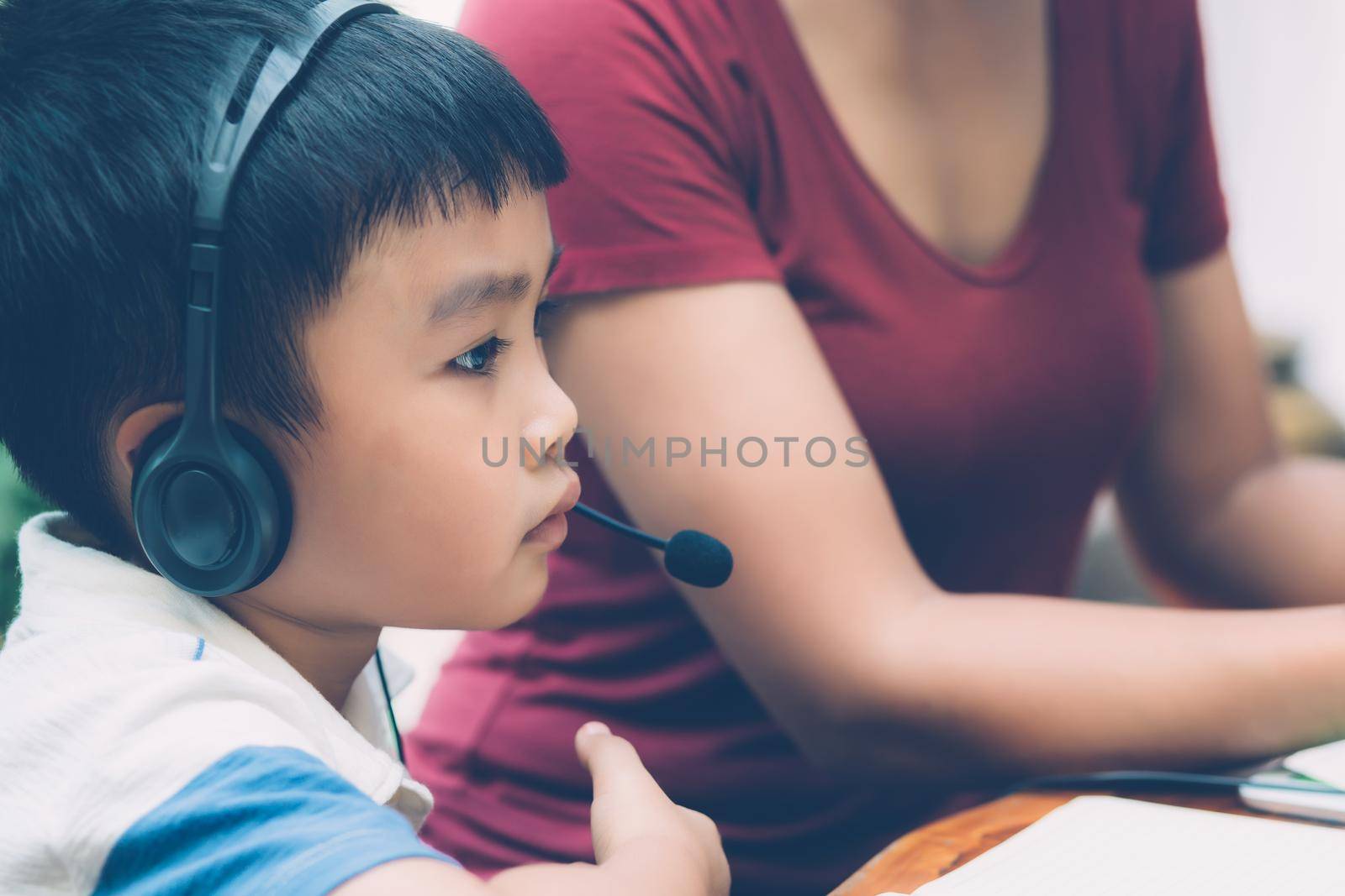 Young asian mother and son using laptop computer for study and learning together at home, boy wearing headphone for e-learning with distancing, teacher or mom support child, education concept.