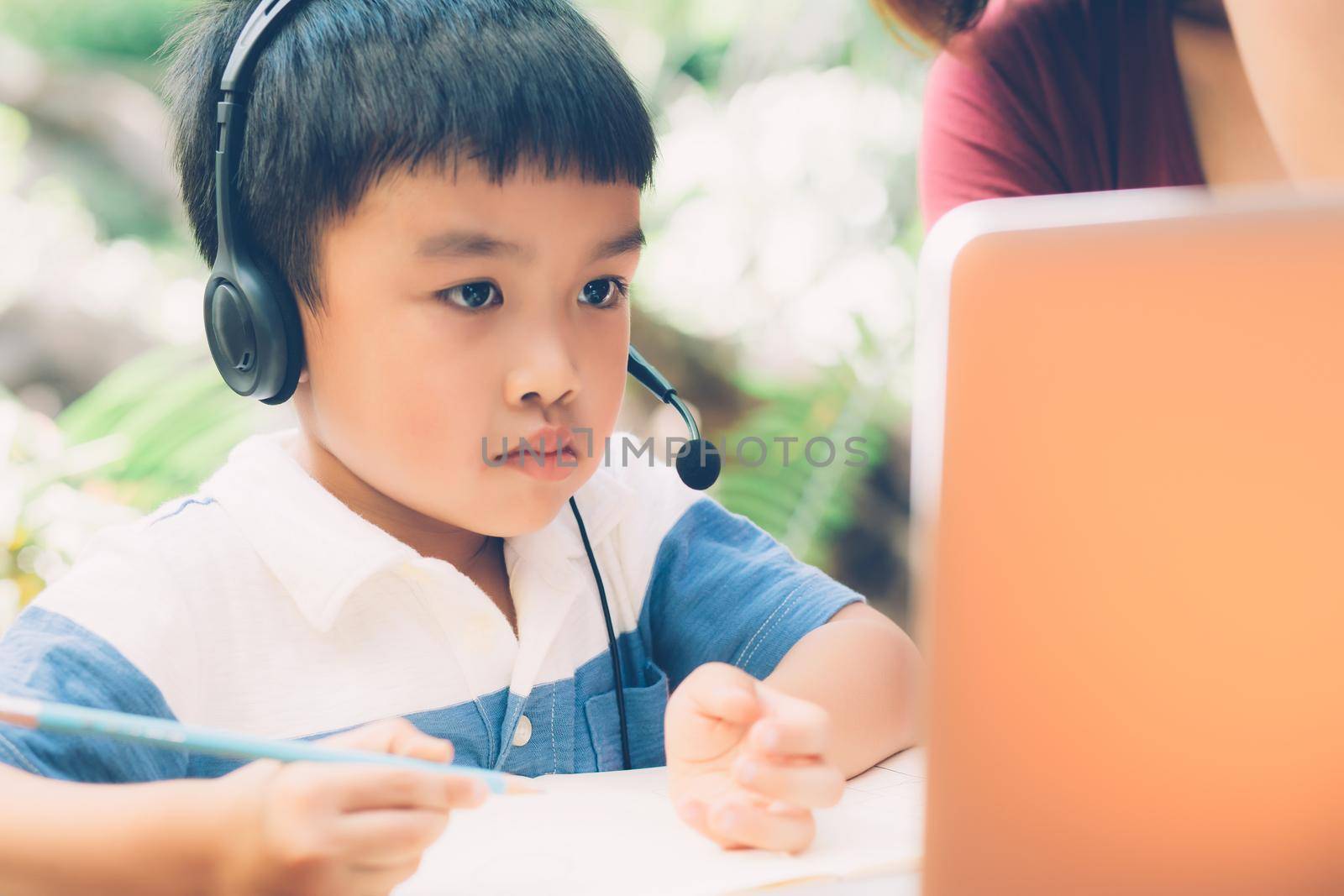 Young asian mother and son using laptop computer for study and learning together at home, boy writing on notebook for homework and wearing headphone, teacher or mom support child, education concept. by nnudoo