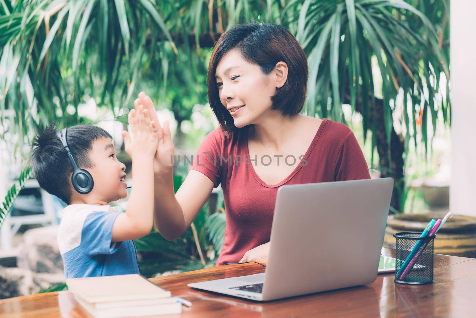 Young asian mother and son using laptop computer for study and learning and hight five together at home, boy wearing headphone for e-learning with distancing, mom support child, education concept. by nnudoo
