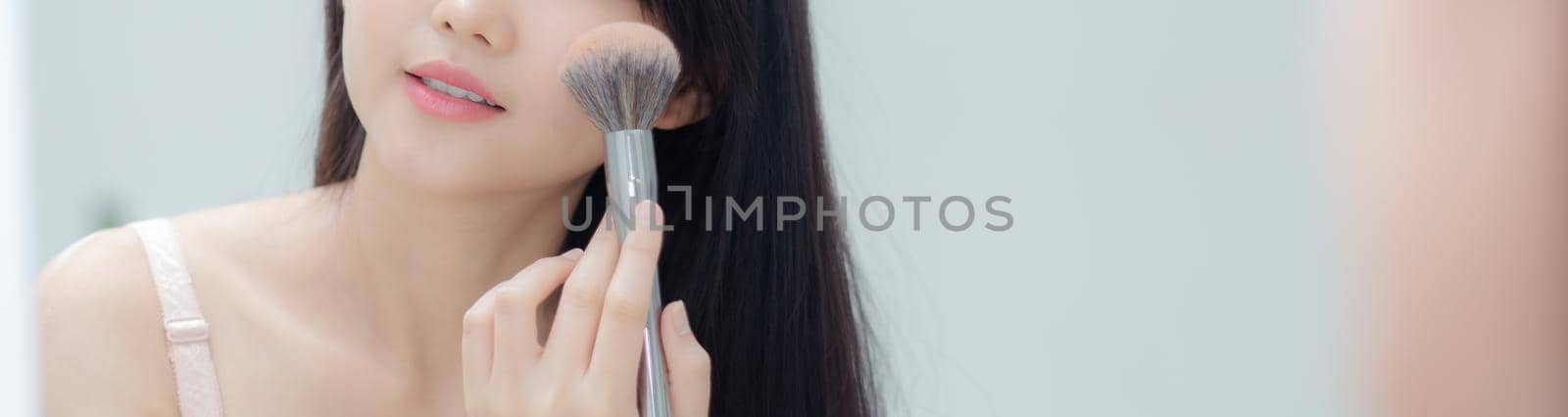 Beauty portrait young asian woman smiling with face looking mirror applying makeup with blusher cheek in the room, beautiful girl blush applying facial, skin care and cosmetic concept, banner website.