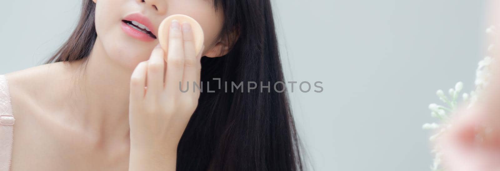 Beautiful young asian woman applying powder puff at cheek makeup of cosmetic looking mirror, beauty of girl with skin face at room, facial having smooth, health and wellness concept, banner website.
