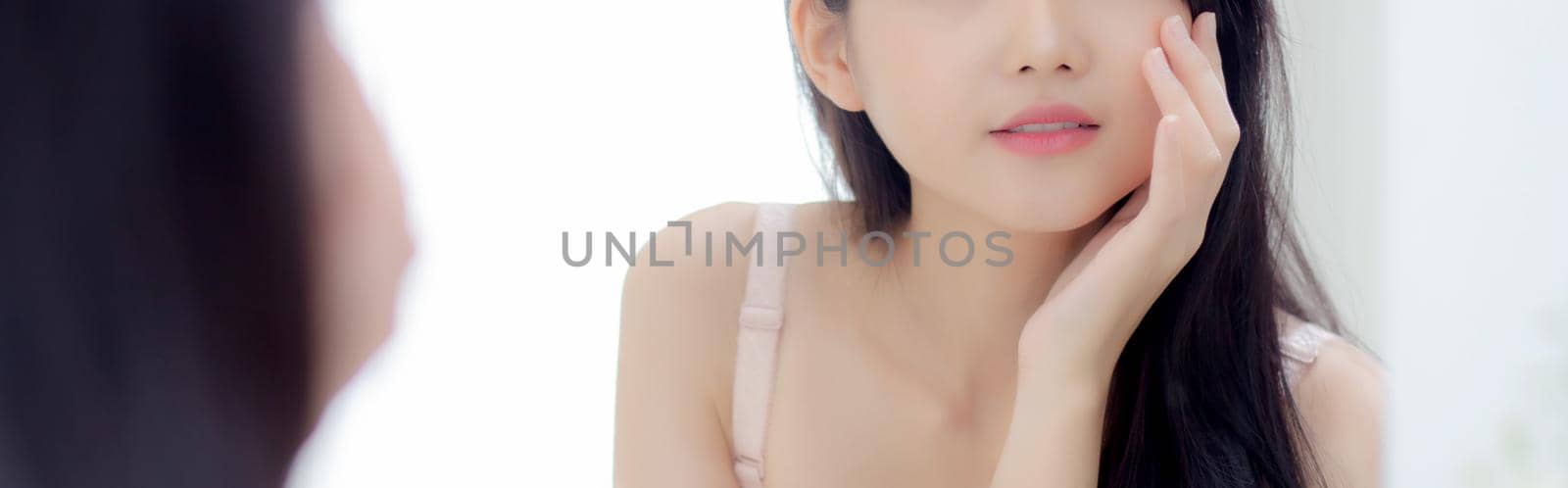 Beautiful young asian woman look at mirror of checking face with skin care and cosmetic for hygiene, beauty girl happy clean facial with cream or lotion and for health, banner website.