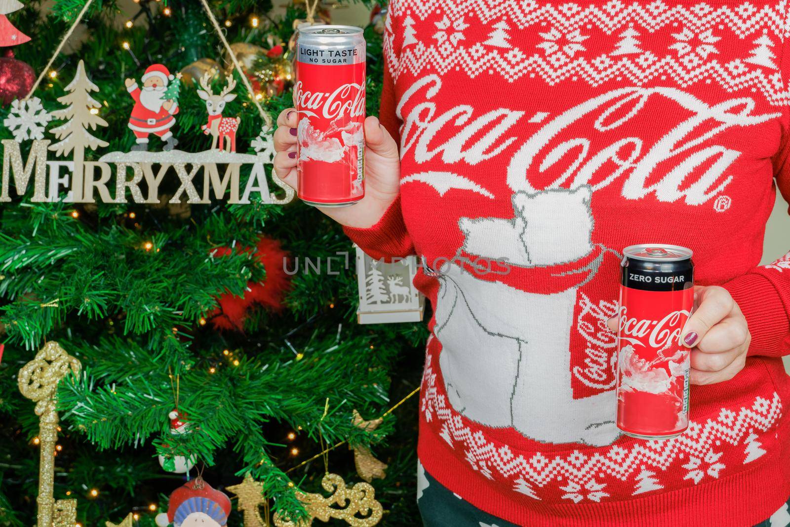 Woman wearing sweater with logo displays seasonal beverage before decorated tree.