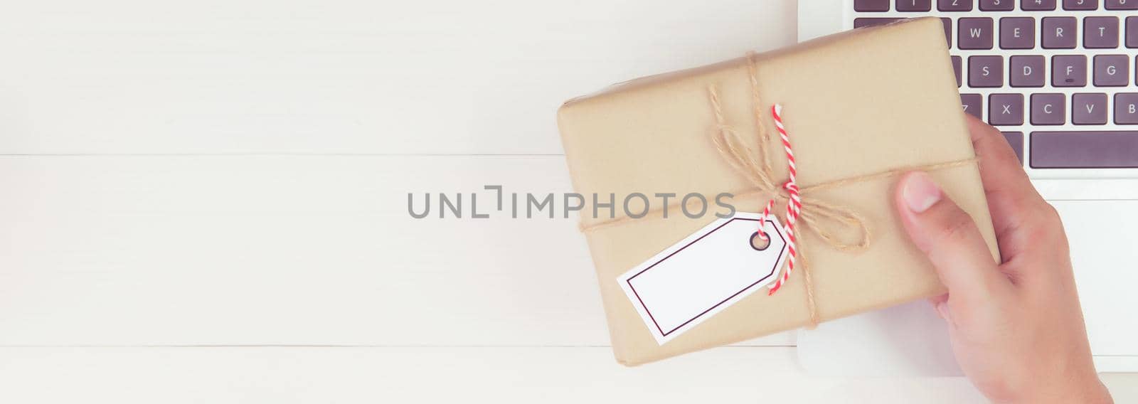 Hand of man holding gift box and tag having laptop computer on wooden table, present giving for Christmas day or birthday, season and celebrate, top view, flat lay, online shopping concept, banner website.