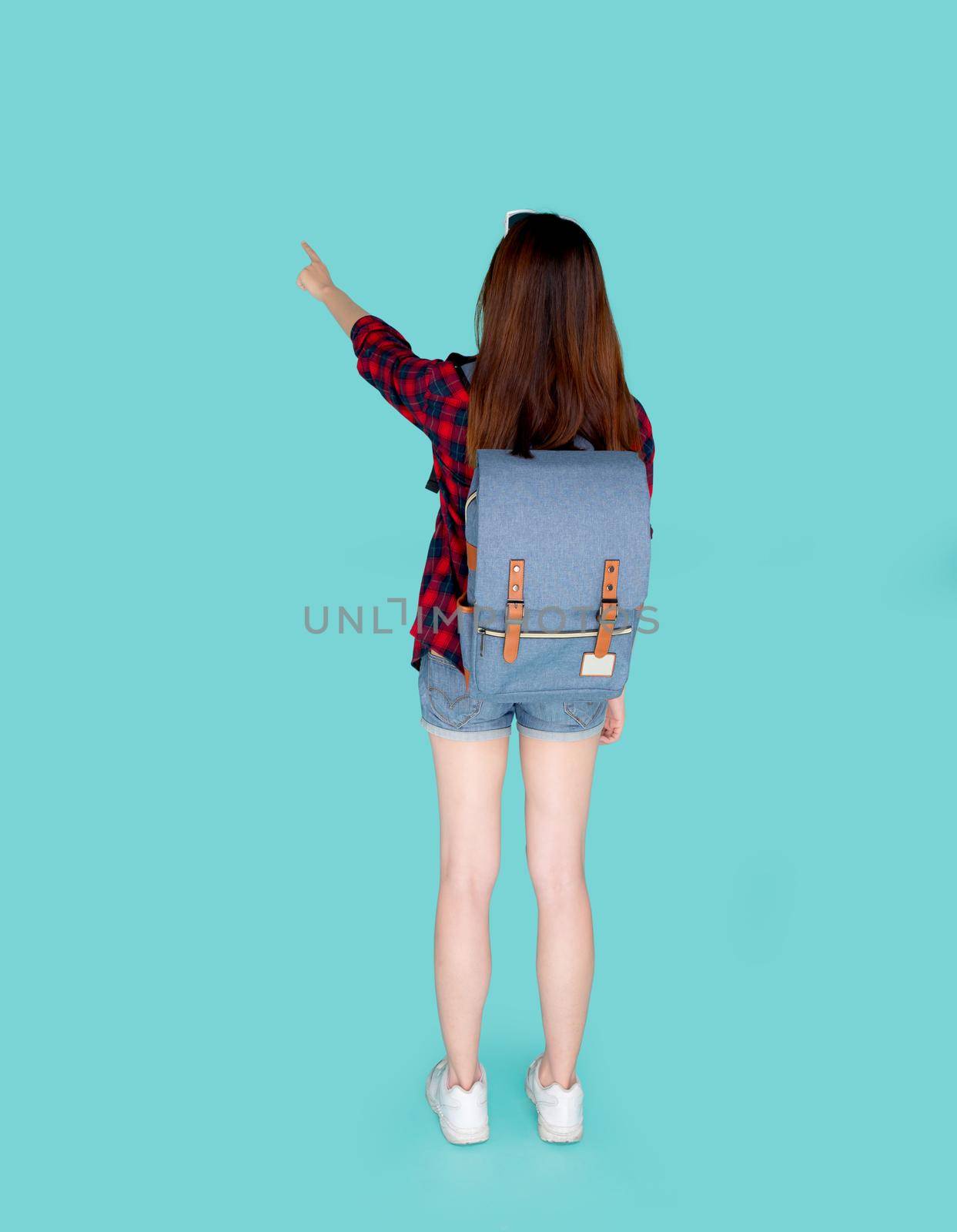 Back view of beautiful young asian woman travel with backpack pointing something isolated on blue background, rear asia girl is student or tourist in summer trip, holiday concept.