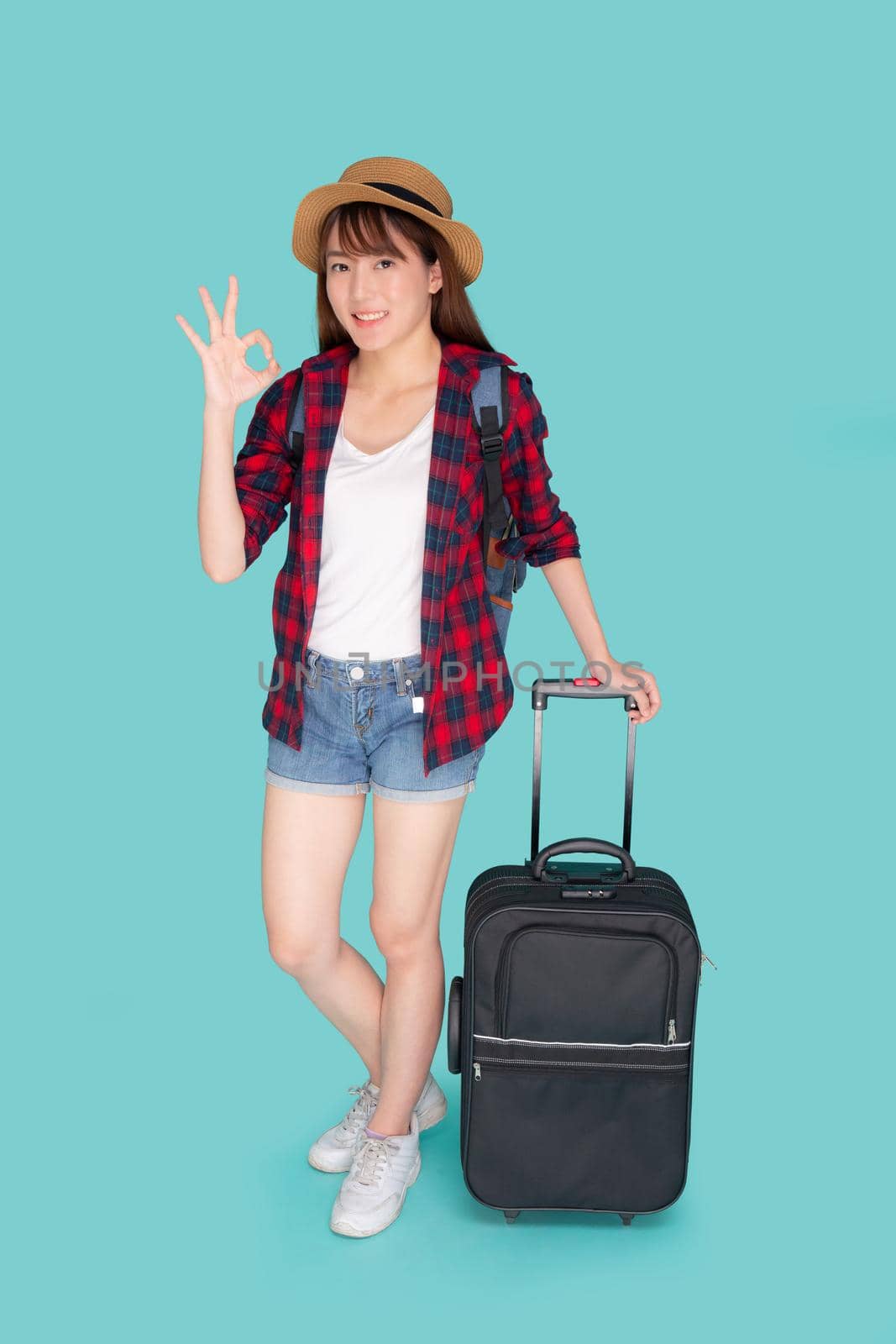 Beautiful young asian woman pulling suitcase isolated on blue background, asia girl cheerful holding luggage walking and gesture ok in vacation with excited, journey and travel concept.