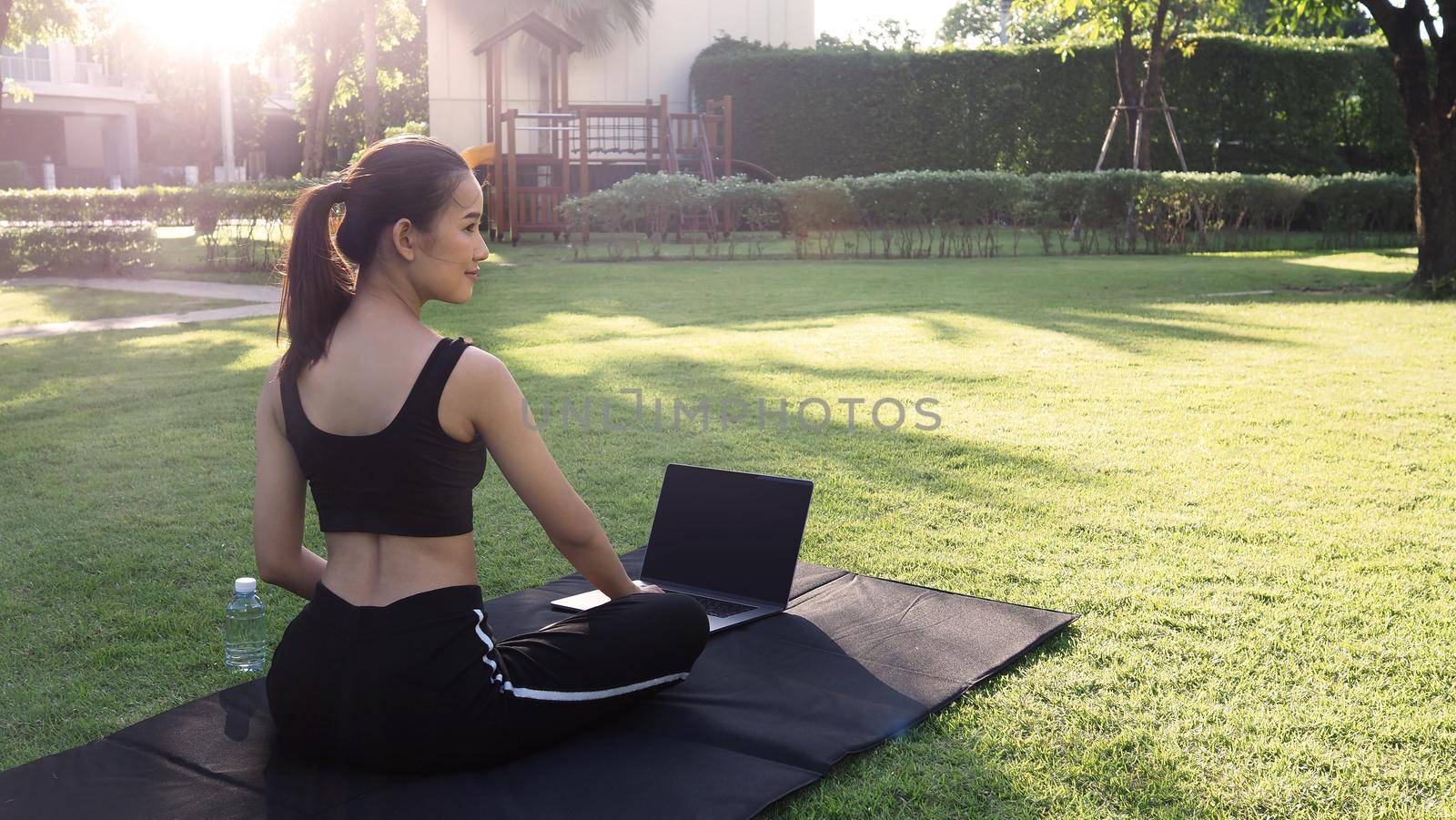 A sporty woman or online instructor in sportswear is sitting and practicing yoga on the mat with computer notebook and a bottle of water and is using a laptop at home in the backyard. Sport and recreation concept.