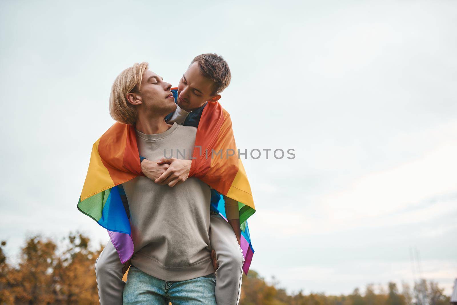 Romantic gay couple hugging, kissing and holding hands outdoors. Two handsome men holding LGBT pride flag.