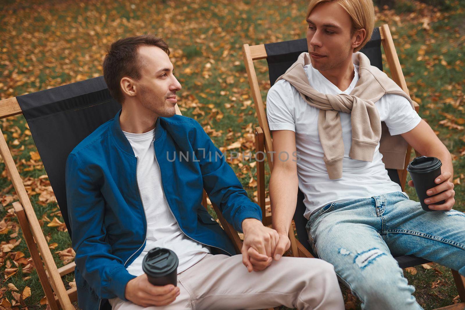 Loving gay couple sitting outdoors together and holding hands while drinking coffee. Two handsome men having romantic date in park. LGBT concept.