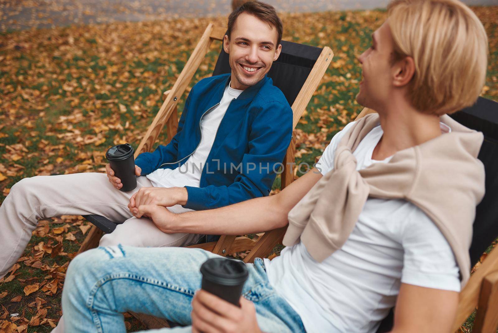 Loving gay couple sitting outdoors together and holding hands while drinking coffee. Two handsome men having romantic date in park. LGBT concept.